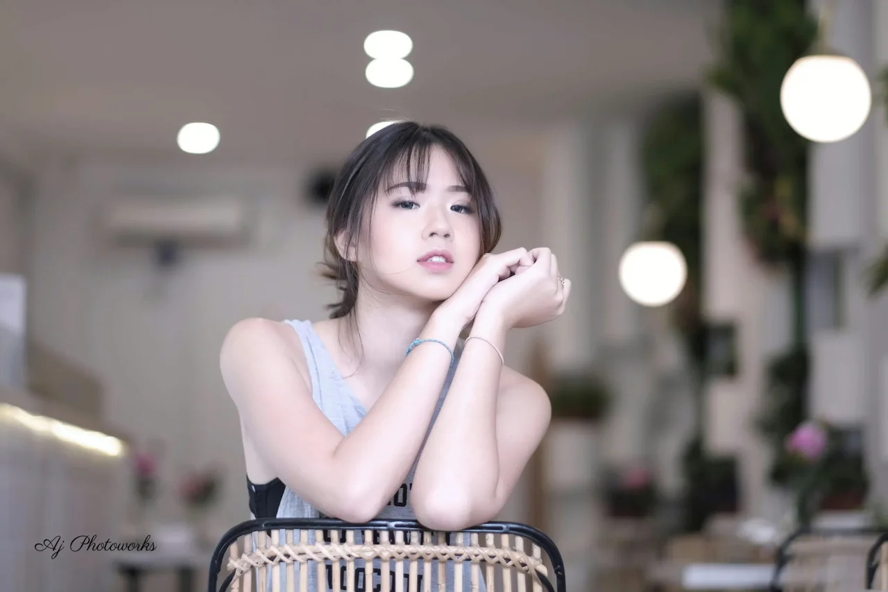 A young woman rests her arms on a wicker chair in a softly lit cafe.