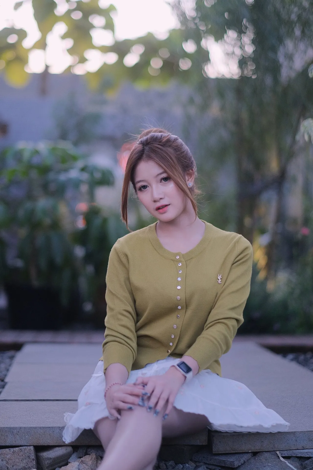 A young woman sits outdoors near foliage, casting a gentle, soft gaze toward the viewer.