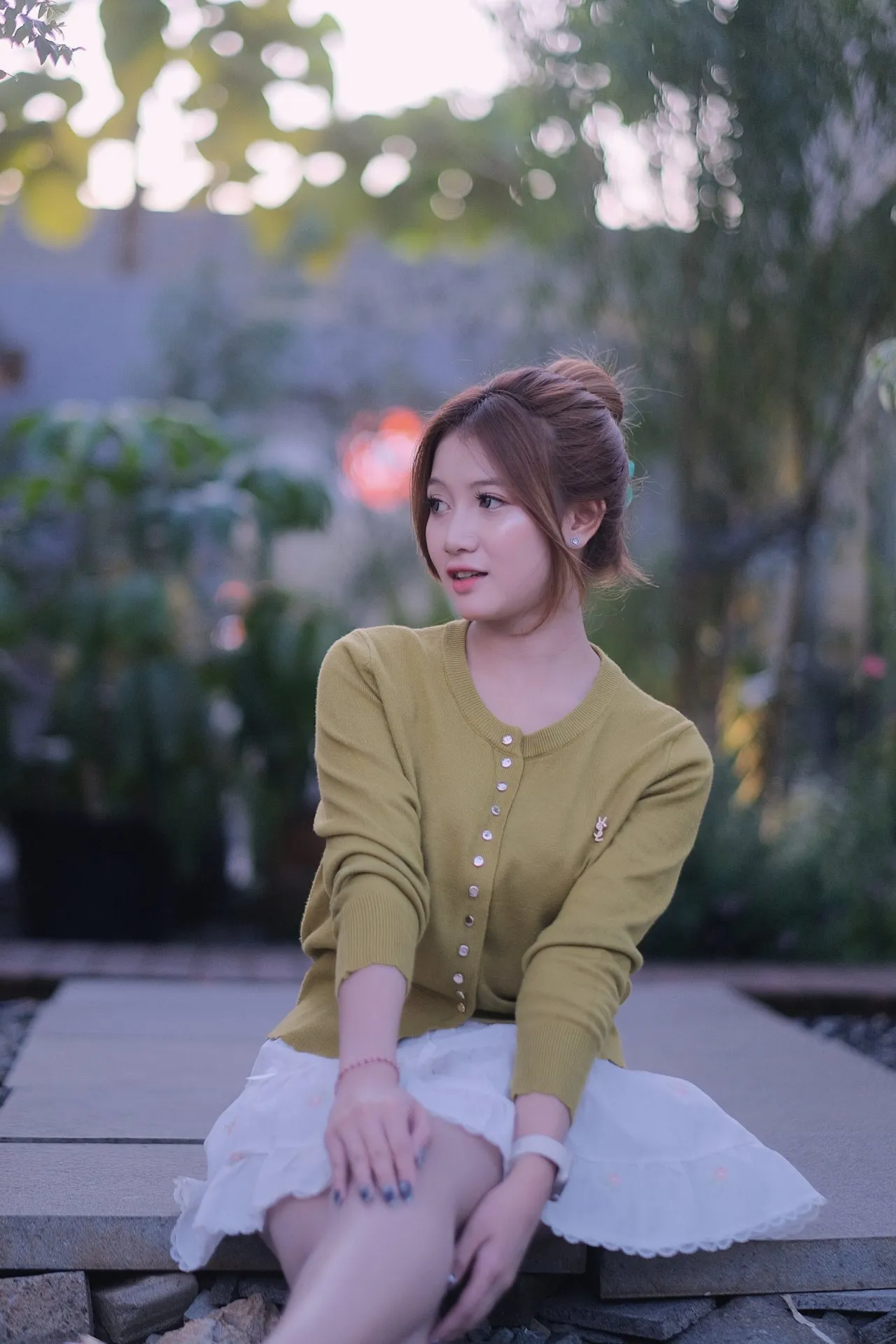 A thoughtful young woman sits outdoors among soft bokeh lights in a serene, muted setting.