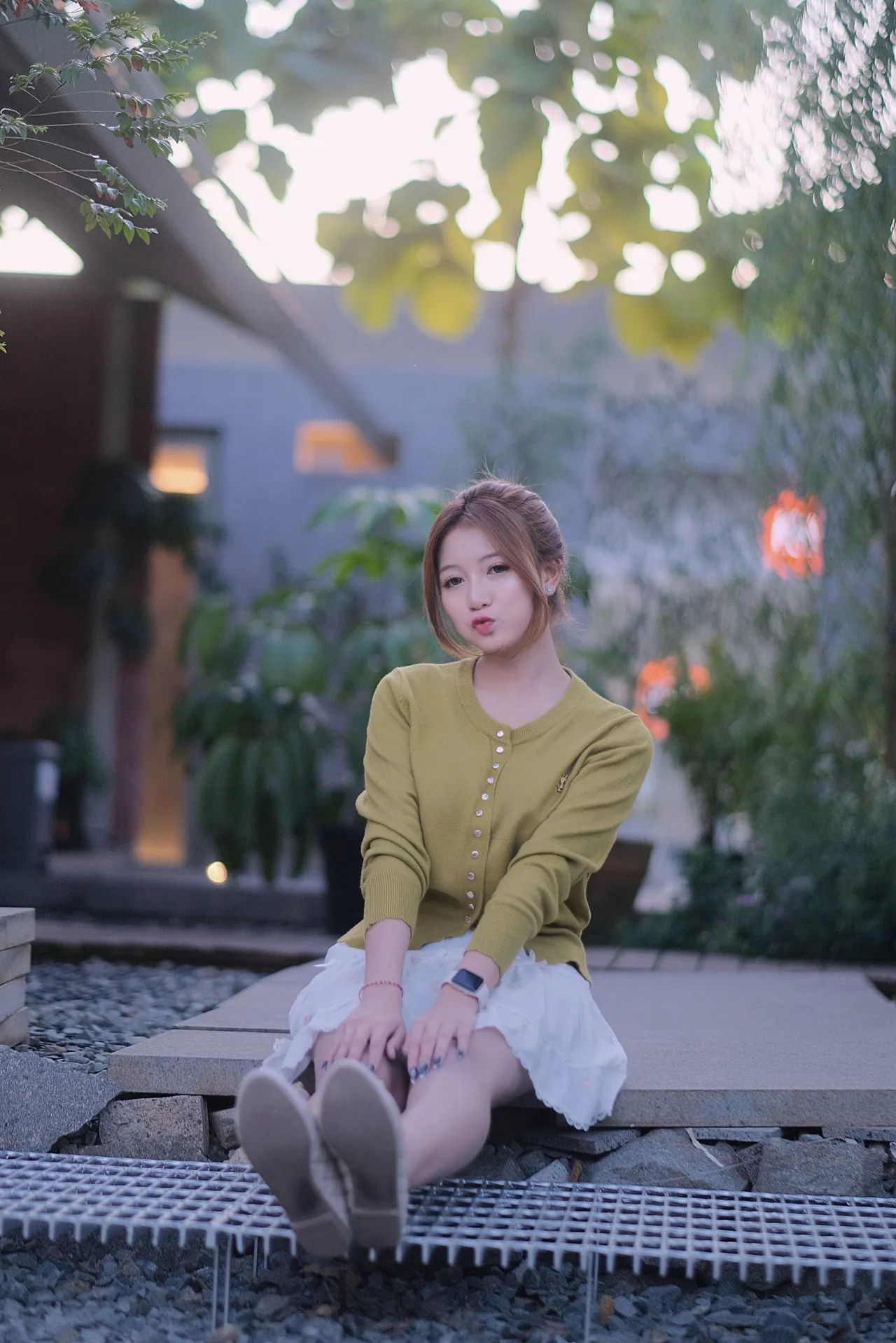 A young woman poses subtly outdoors among lush greenery with a soft, hazy light.